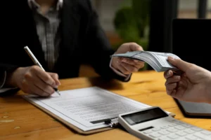 Pessoa assinando contrato enquanto recebe uma pilha de dinheiro em uma mesa de escritório, com um dispositivo eletrônico ao lado.