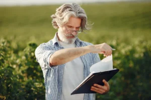 Homem com cabelo grisalho, barba e óculos, segurando uma prancheta e analisando notas ou dados em um campo agrícola sob luz natural.