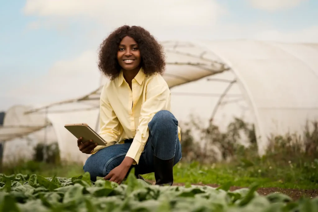 Jovem mulher sorridente usando celular, em campo com estufas ao fundo, promovendo agricultura sustentável e inovação agrícola.
