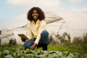 Jovem mulher sorridente usando celular, em campo com estufas ao fundo, promovendo agricultura sustentável e inovação agrícola.