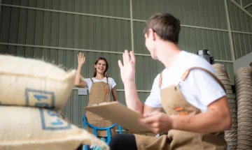 Imagem de jovens trabalhadores em revenda agricola sorrindo e acenando um para o outro enquanto seguram prancheta e estao cercados por sacos de material