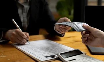 Pessoa assinando contrato enquanto recebe uma pilha de dinheiro em uma mesa de escritório, com um dispositivo eletrônico ao lado.
