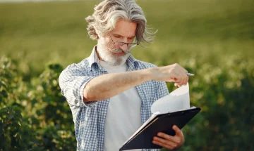 Homem com cabelo grisalho, barba e óculos, segurando uma prancheta e analisando notas ou dados em um campo agrícola sob luz natural.