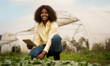 Jovem mulher sorridente usando celular, em campo com estufas ao fundo, promovendo agricultura sustentável e inovação agrícola.