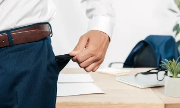 Pessoa com terno ajustando o bolso de uma calça social, ambiente de escritório com mesa, papéis, mochila, óculos e planta ao fundo.